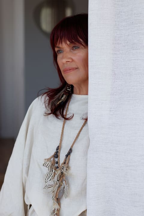 Dawn in cream linen beside a sheer curtain, gazing out in quiet reflection